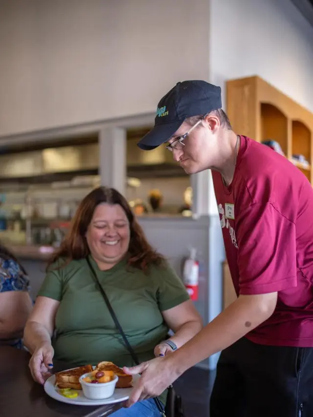 woman in green shirt sits at table where server is serving her a plate of food