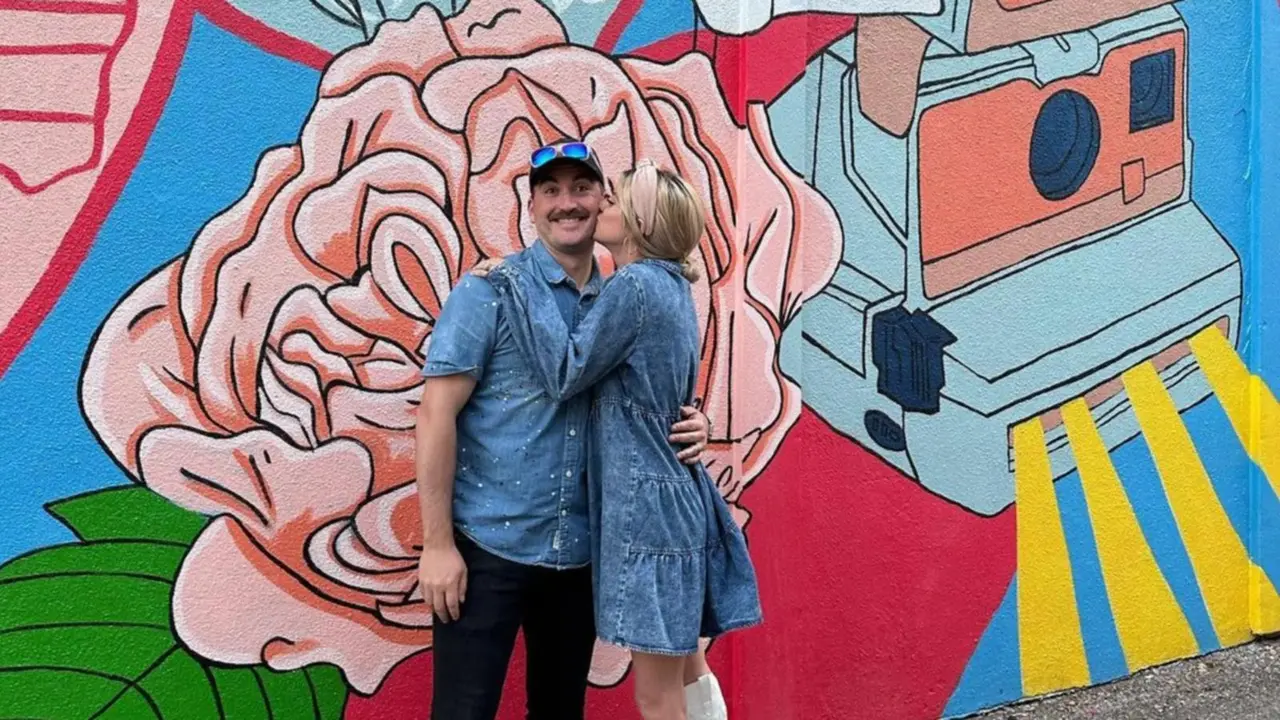 A young blonde woman in a denim dress and white boots kisses a young mustached man on the cheek in front of a colorful mural