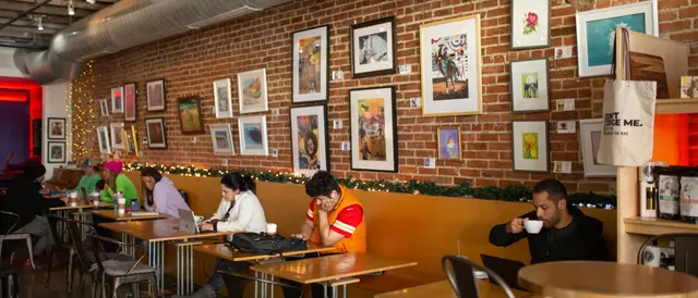 row of small tables with people sitting, drinking coffee and looking at phones or laptops