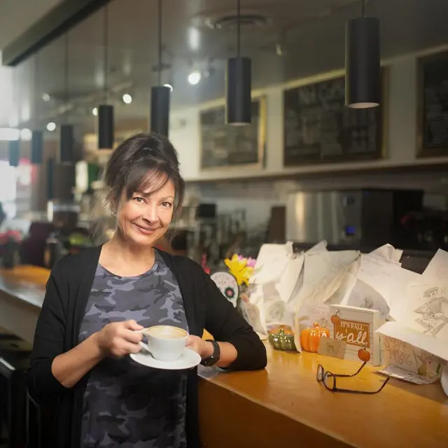 A smiling brunette woman standing at a counter holding a coffee cup