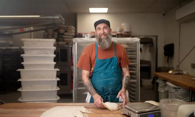 Photo of a bearded man in a rust-colored tee shirt and blue apron, working with bread dough on a big cutting board