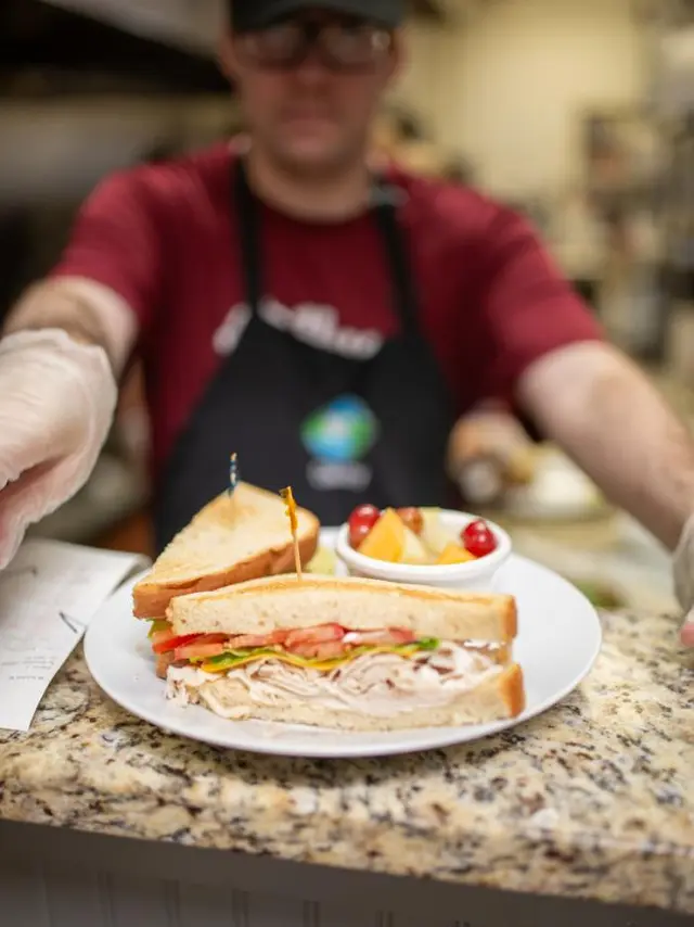 a plate with a sandwich and cup of fruit sitting on a counter with person in the background