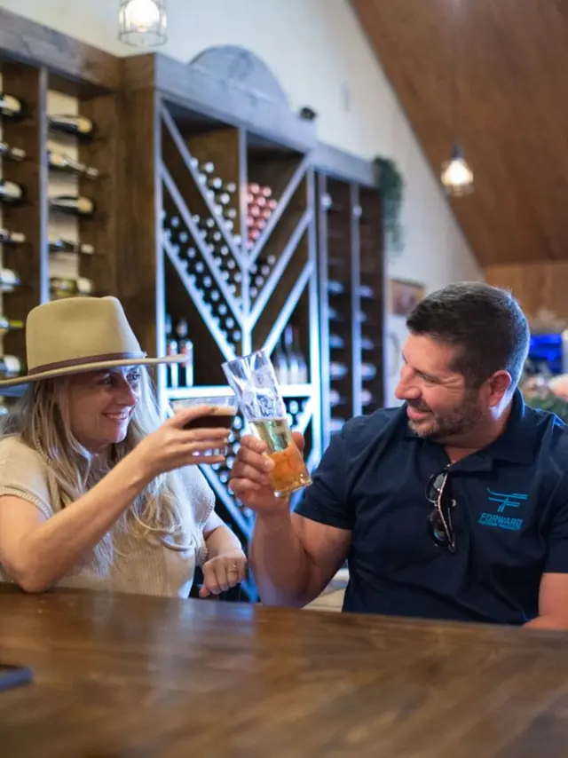 A man and a woman in a hat toasting at the bar