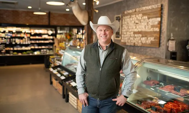 Man in a cowboy hat, long sleeves, and a zippered vest standing in front of a meat counter full of cuts of beef
