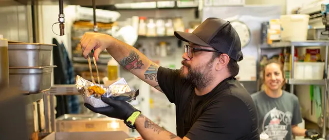 bearded man in ball cap with tongs getting food out of a big pot on stove