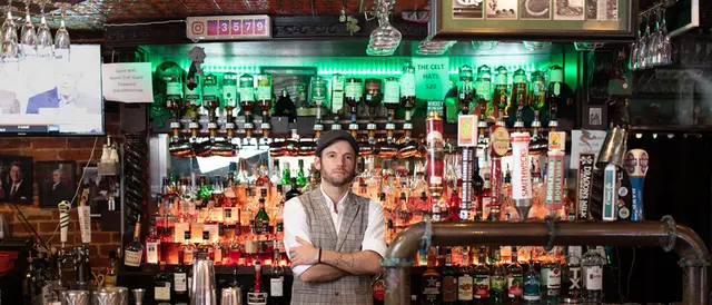 Man in a vest and driving hat stands, arms folded, behind a fully-stocked bar