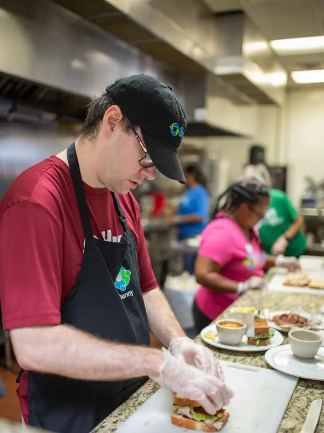 man in glasses, burgundy shirt and black apron prepares food in the kitchen