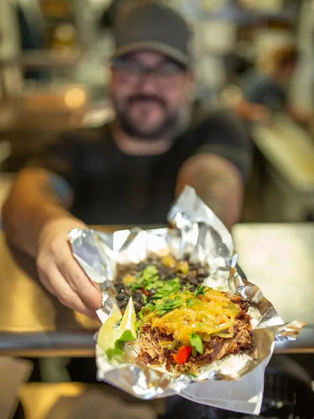 bearded man holding out a street taco in foil